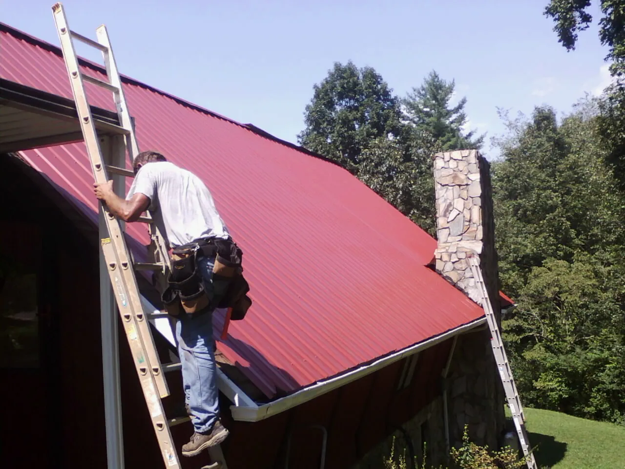 Roofer on a ladder beside a red metal roof and stone chimney — Soffit & Fascia Repair by Roofing Brothers in Cashton